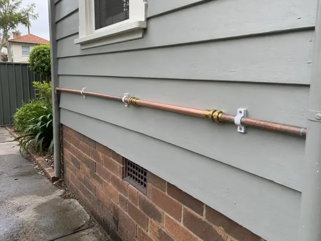 Copper water pipe repaired along an exterior weatherboard wall above brickwork in Mascot